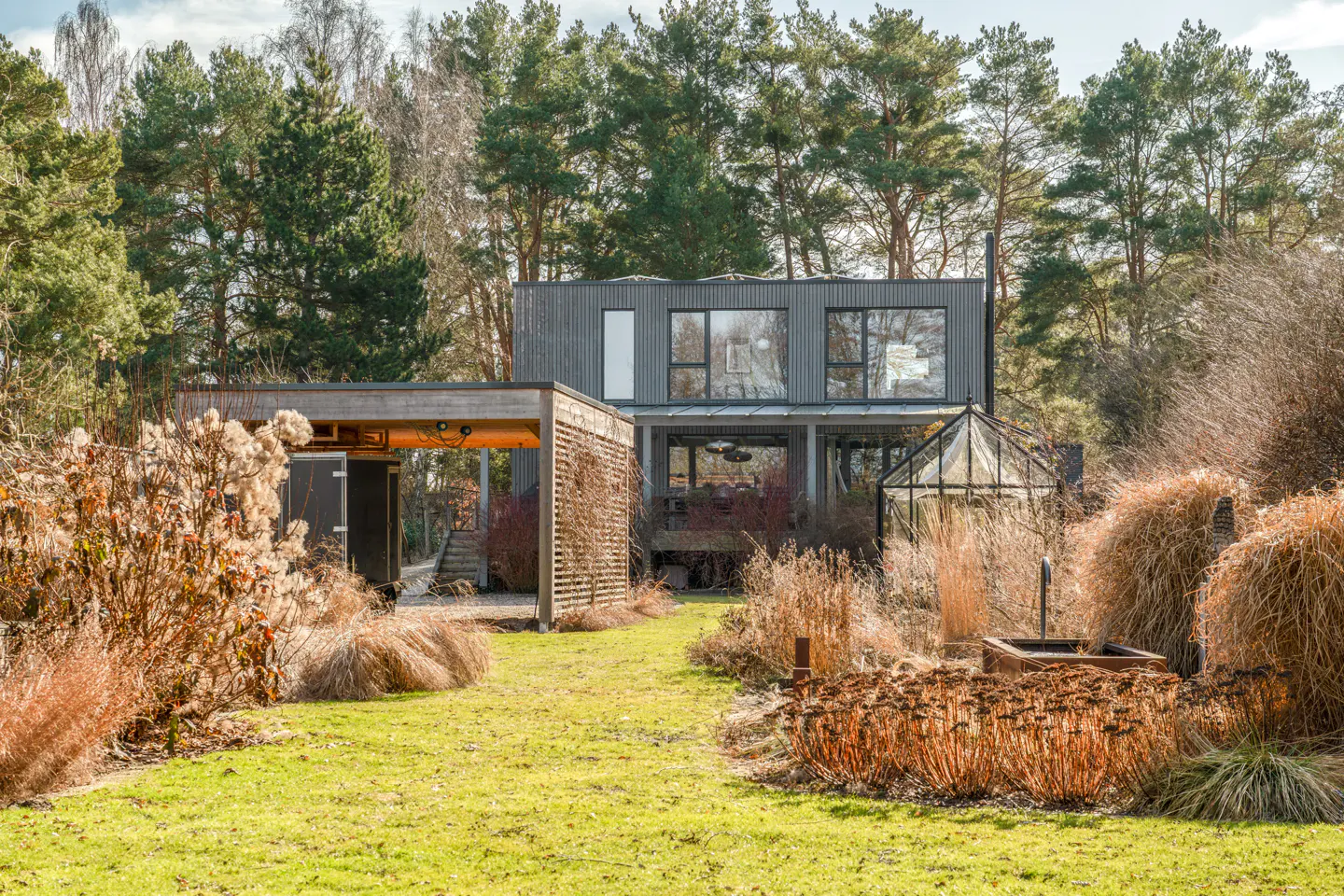 Exterior view of a modern gray house with a greenhouse and a wooden carport surrounded by a green lawn and trees.