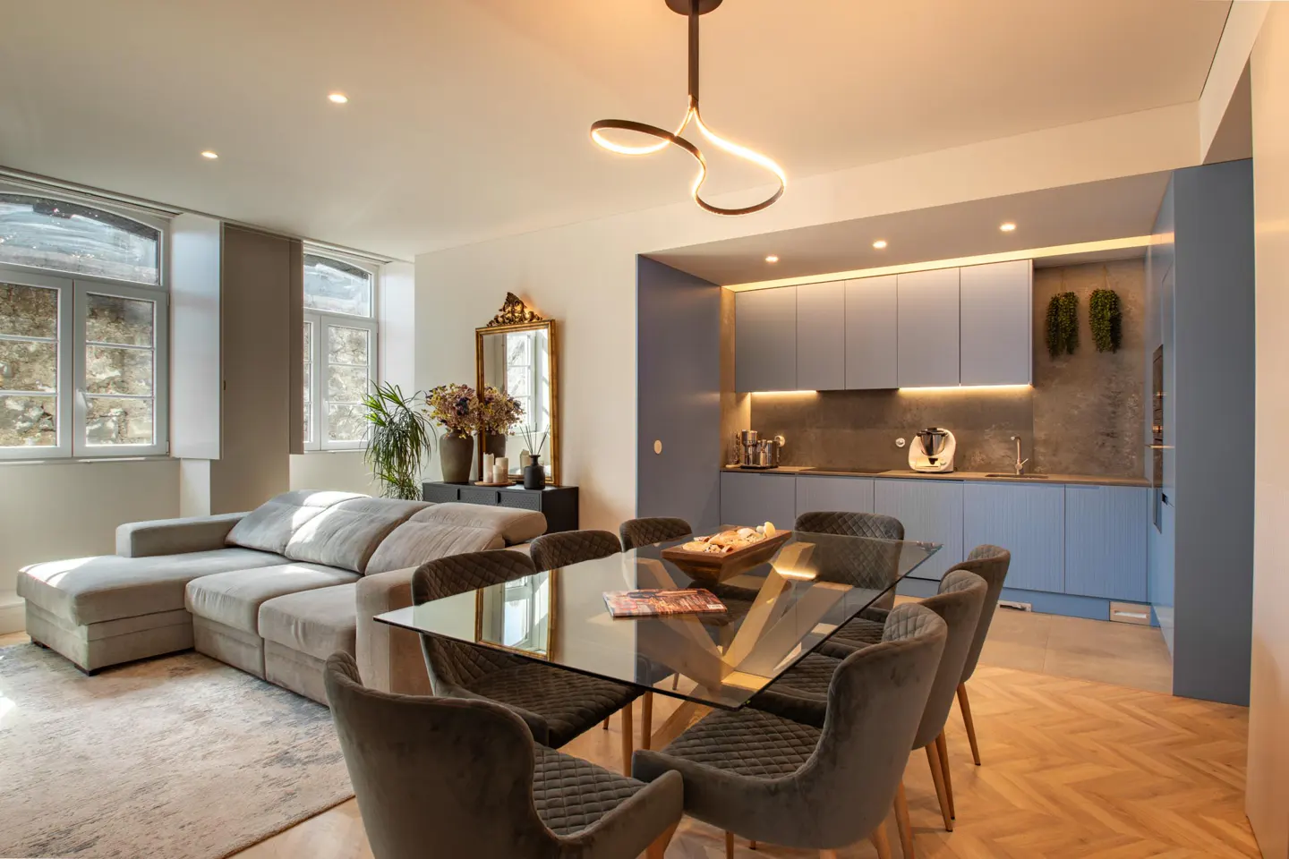 Bright, modern apartment interior with a glass dining table, grey velvet chairs, and a blue kitchen. A beige sectional sofa sits near large windows.