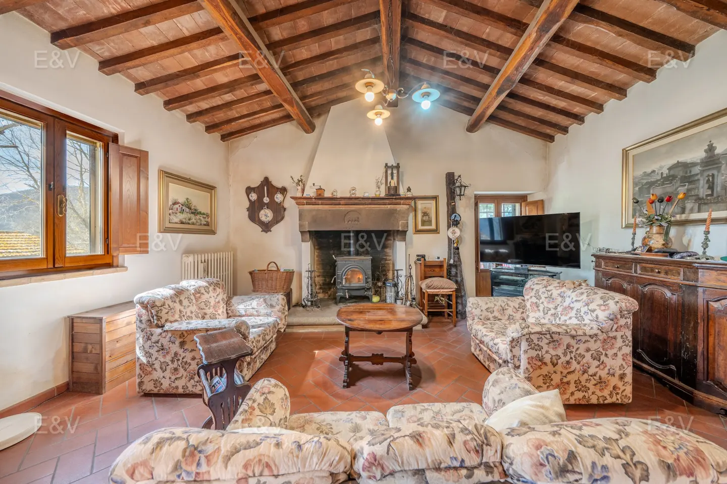 Living room with floral sofas, wood-beamed ceiling, fireplace with wood-burning stove, and terracotta tile floor.