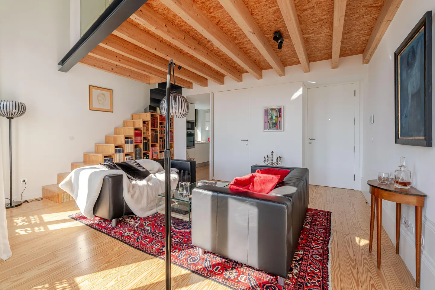 Bright living room with wood floors, black sofas, and a red patterned rug. Stairs with built-in bookshelves lead to a loft.