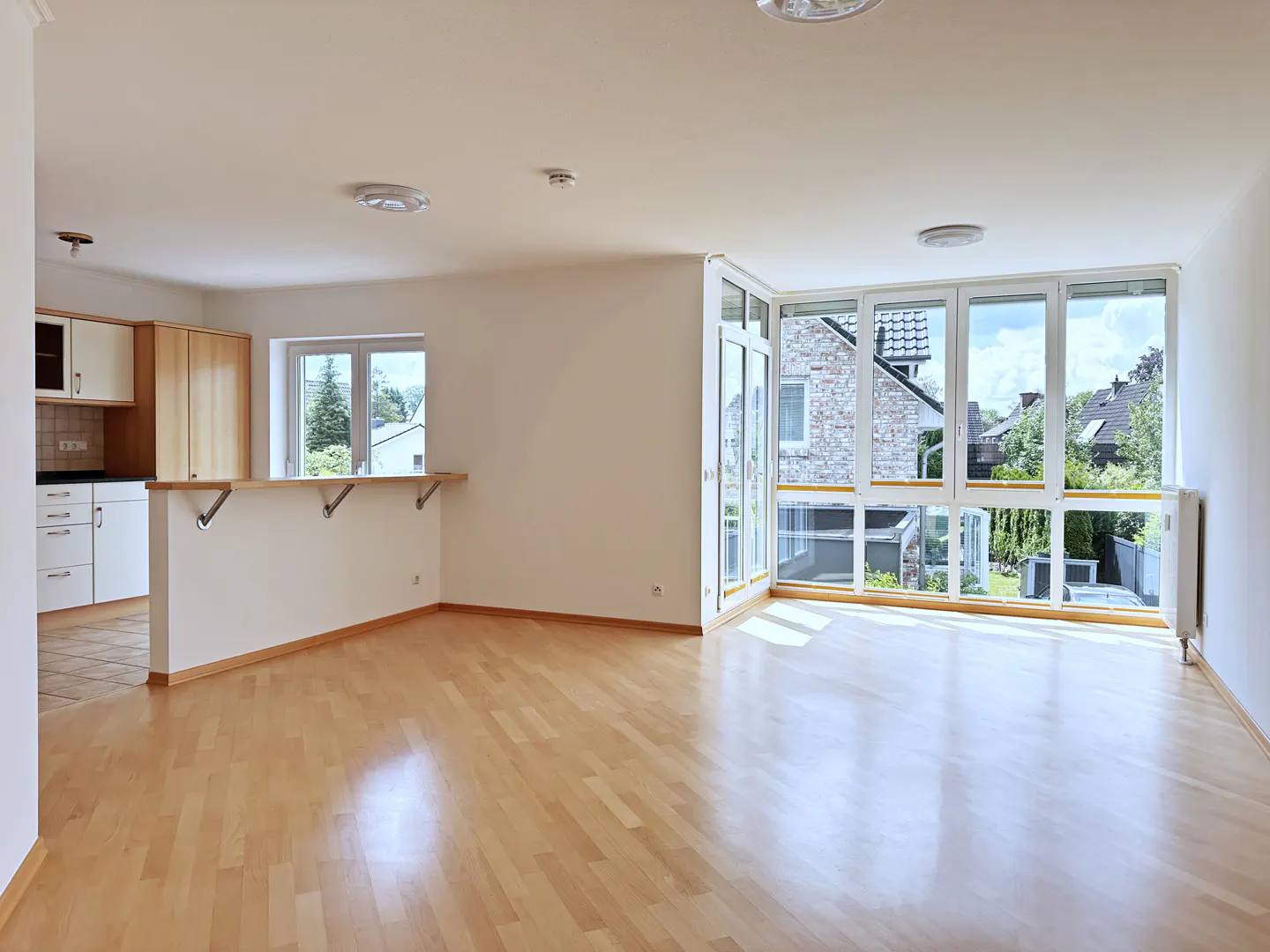 Bright, open-plan living area with light wood floors, white walls, and a kitchen counter overlooking a sunny garden view.