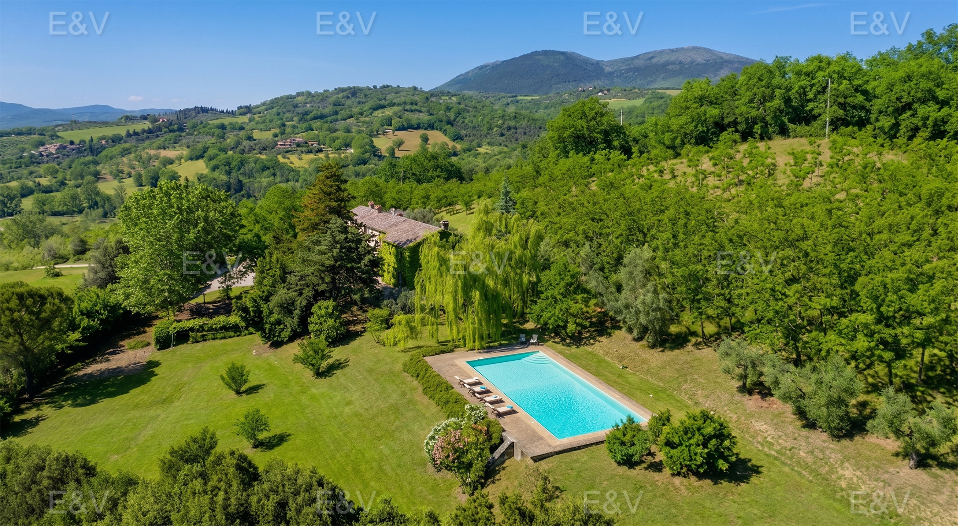 Aerial view of a villa with a pool surrounded by green trees and rolling hills under a blue sky.
