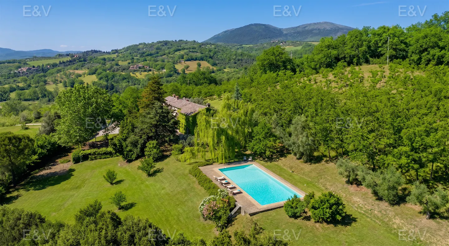 Aerial view of a villa with a pool surrounded by green trees and rolling hills under a blue sky.