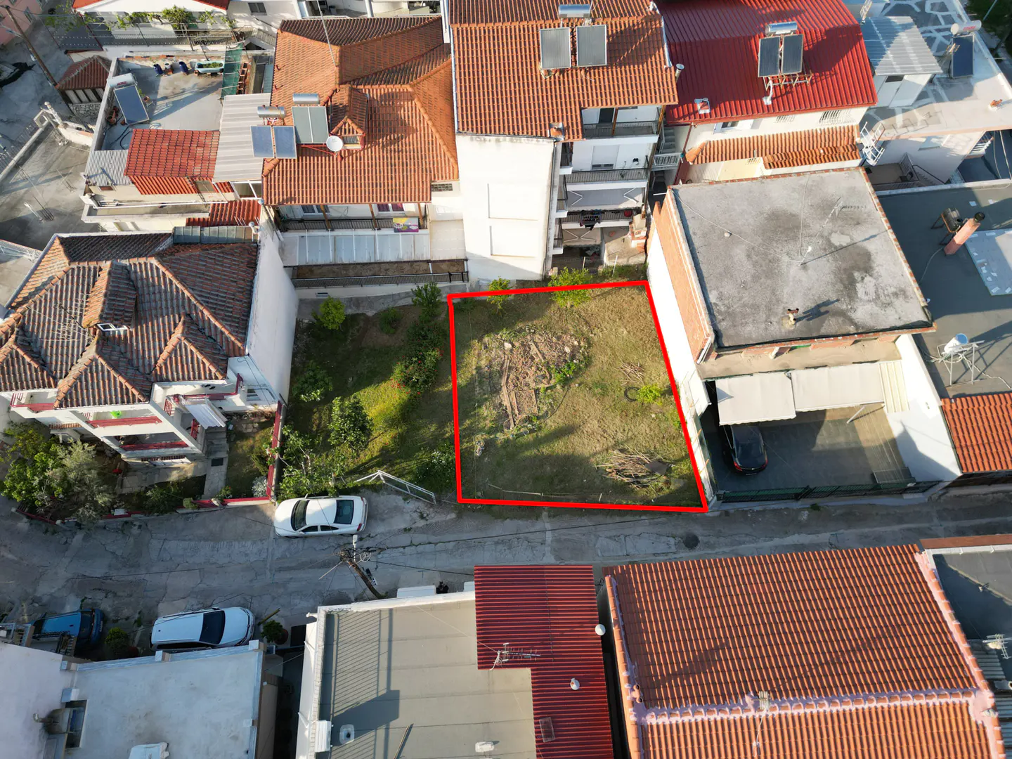 Aerial view of a vacant lot outlined in red, surrounded by houses with orange tile roofs. A street with parked cars is visible.