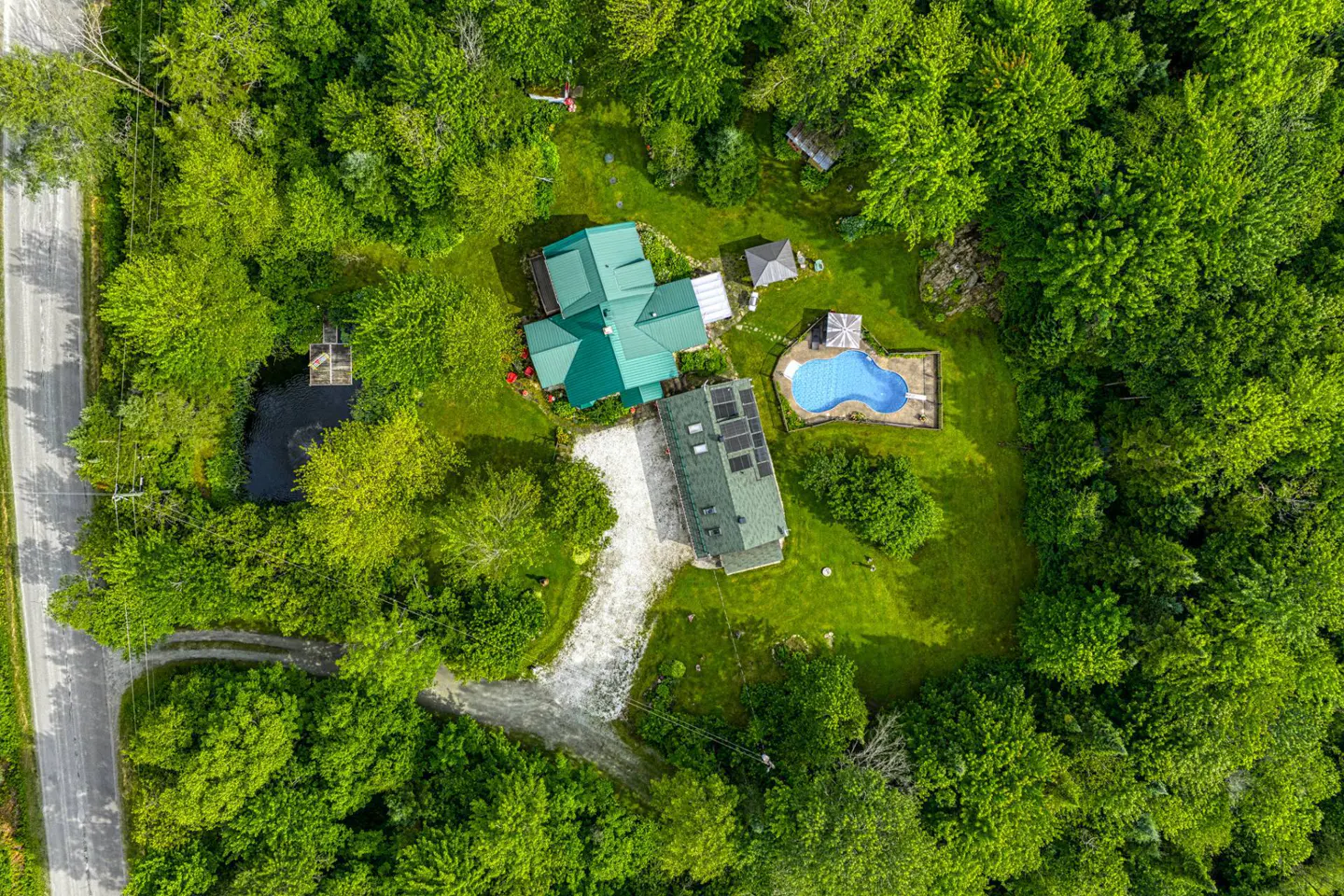 Aerial view of a green-roofed house with a pool, pond, and surrounding lush green trees.