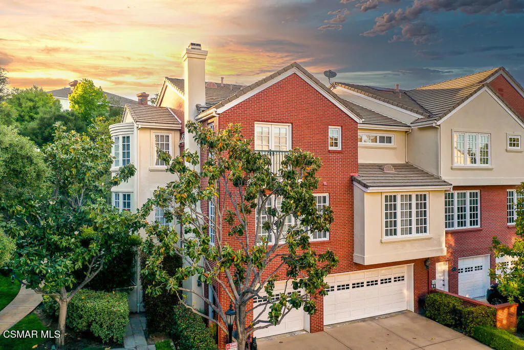 Exterior shot of a multi-story townhouse with a red brick facade and a white garage door, surrounded by green trees.