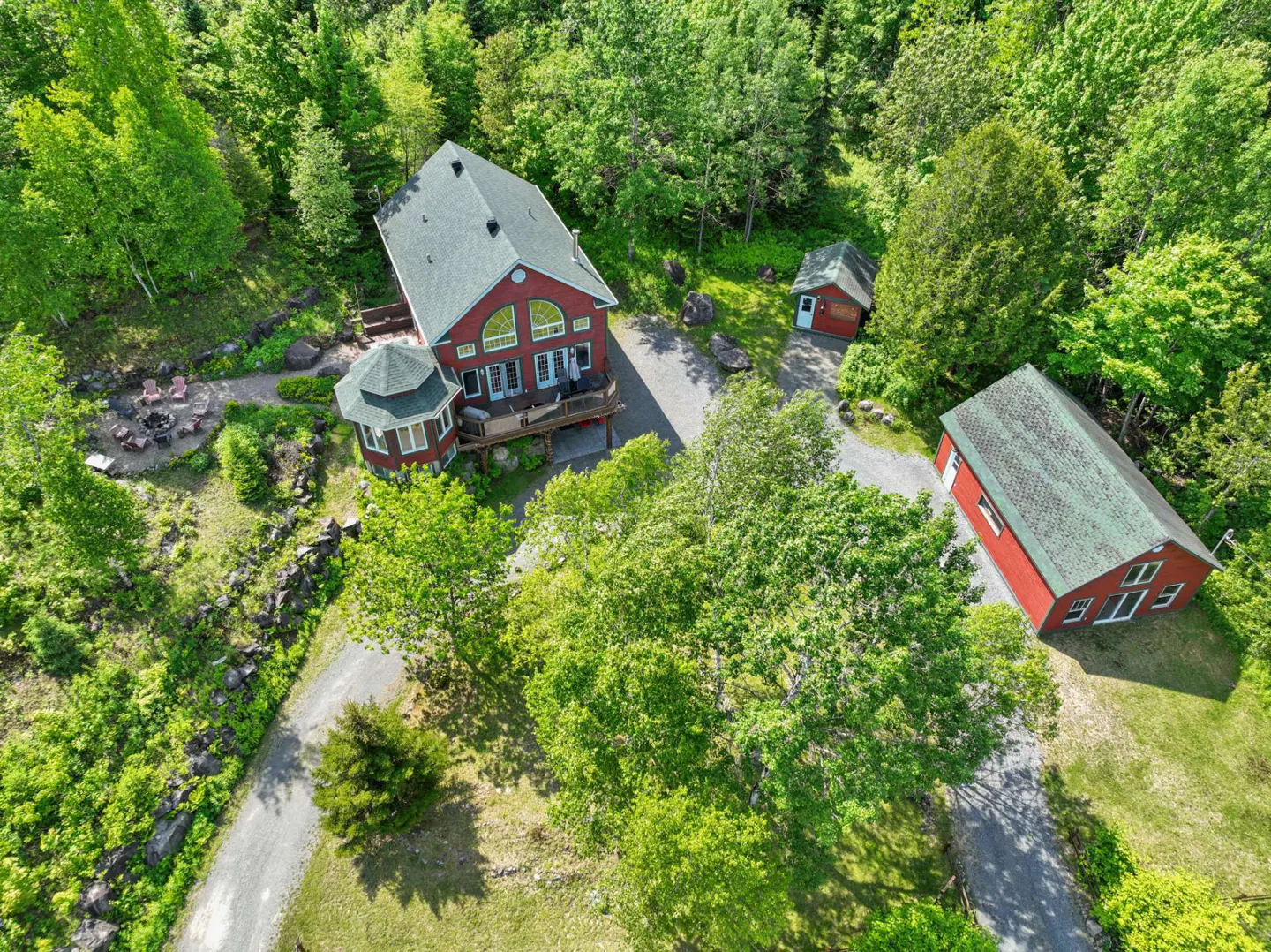 Aerial view of a red house with a green roof, surrounded by lush green trees and a gravel driveway.