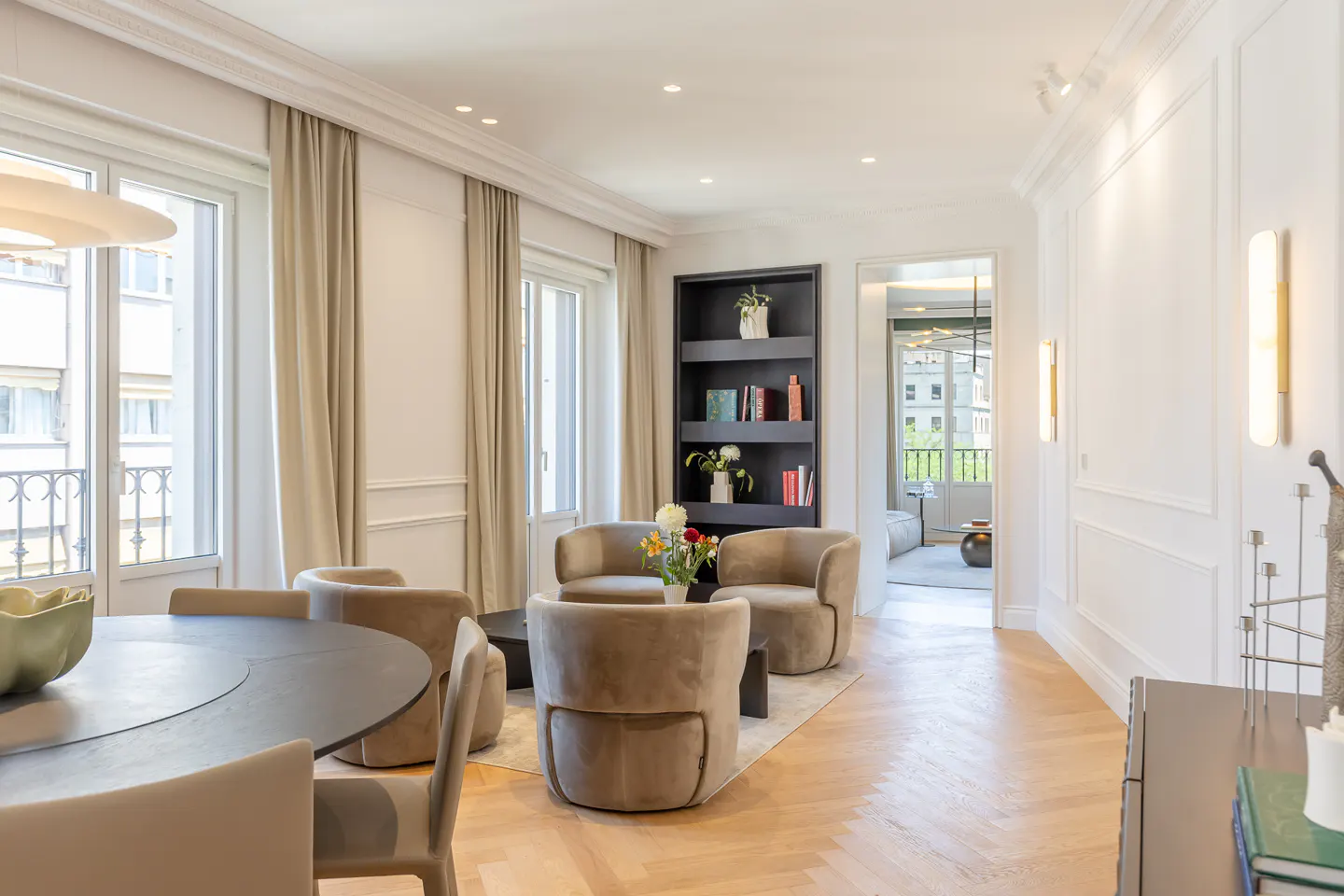 Bright living room with herringbone wood floors, beige armchairs, a black bookshelf, and a dining area with a round table.
