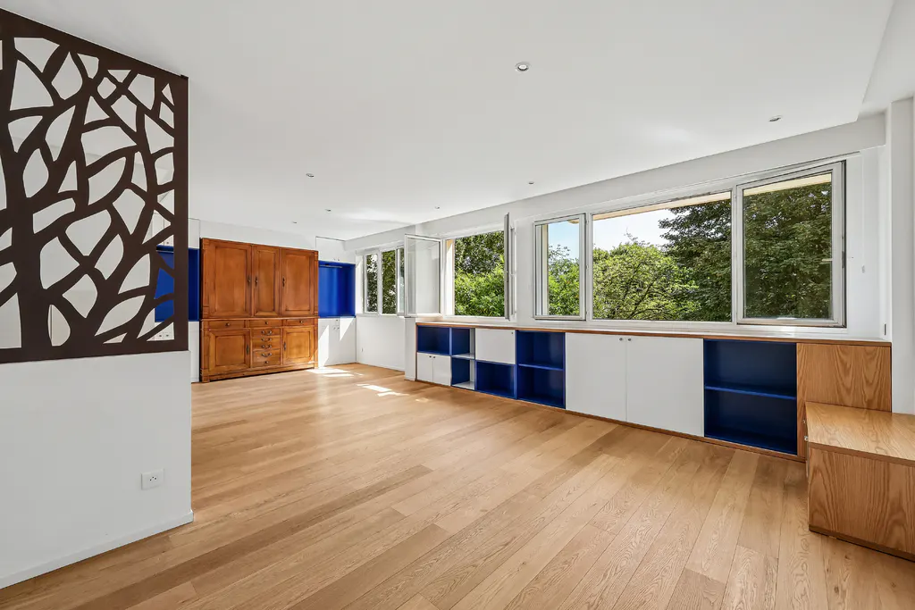 Bright, empty room with wood floors, white walls, and built-in blue and white cabinets under windows with green tree views. A wood cabinet and decorative screen are on the left.