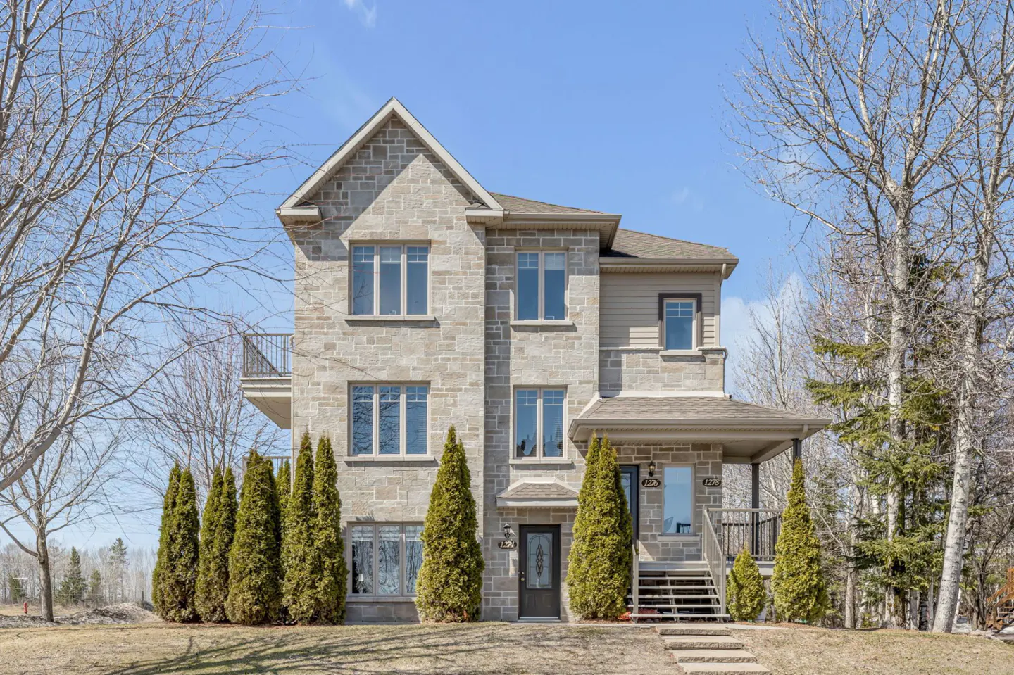Two-story stone house with a balcony, front porch, and evergreen trees in front.