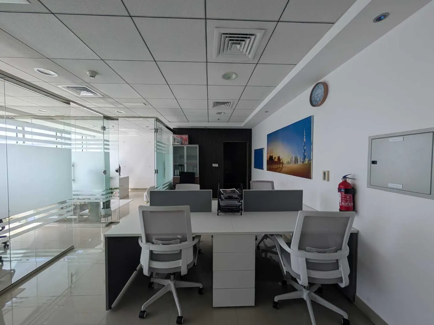 Bright office space with glass-walled rooms, white desks, and gray chairs. A Dubai skyline art piece hangs on the wall.