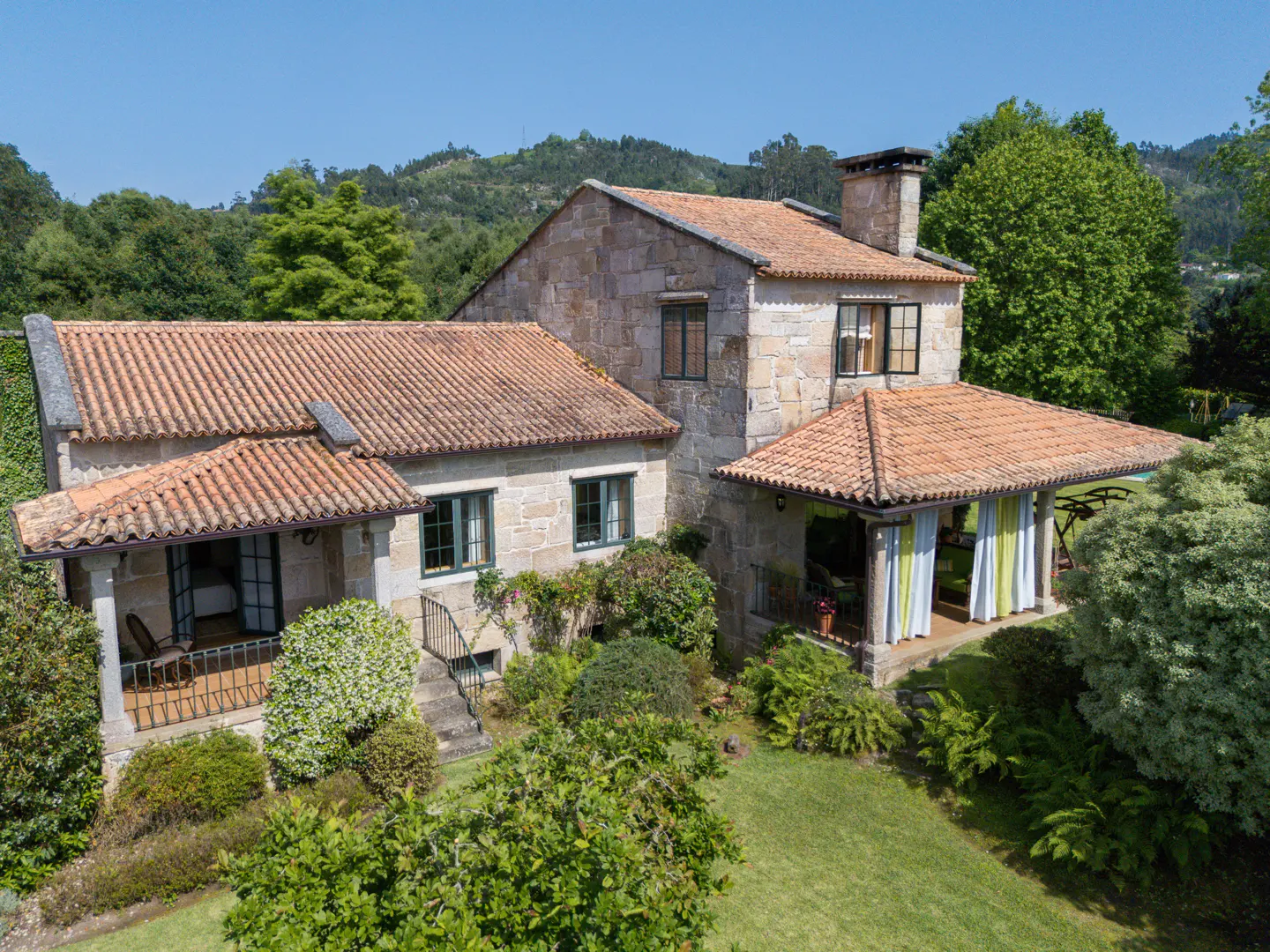 Stone house with a red tile roof, green trim, and a chimney, surrounded by lush green trees and grass.