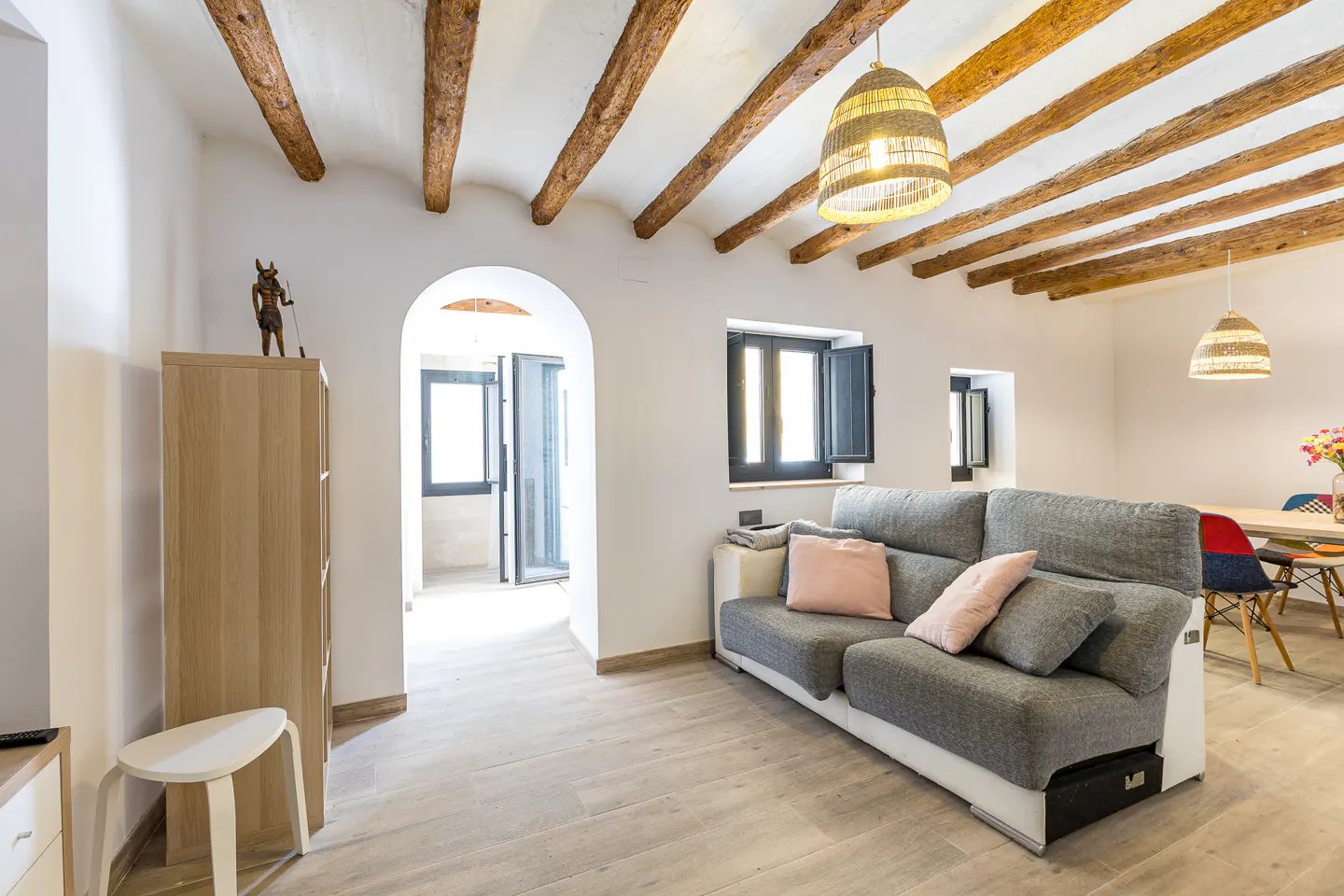 Living room with exposed wooden ceiling beams, a grey sofa with pink pillows, and an arched doorway leading to a hallway.