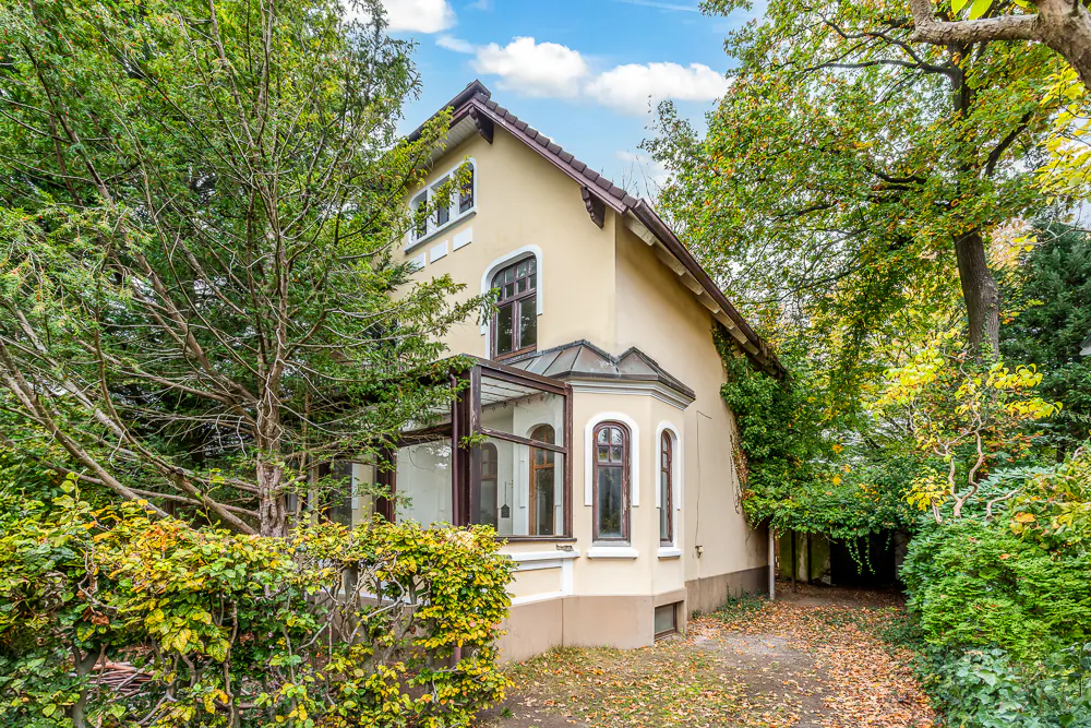 Two-story beige house with brown trim, surrounded by green trees and bushes under a blue sky.