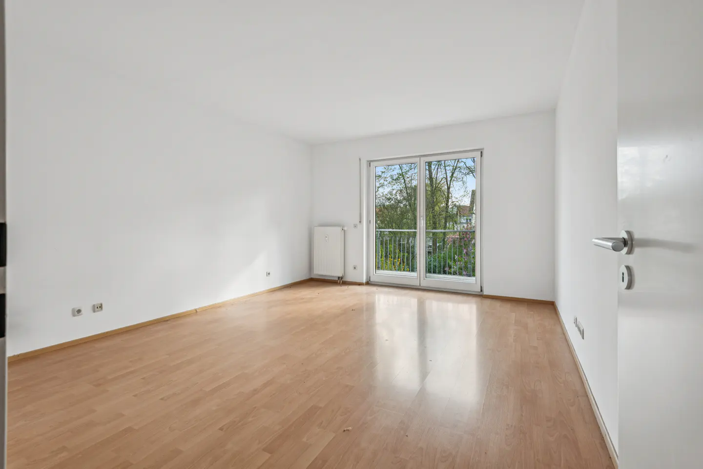 Bright, empty room with light wood floors and white walls. Balcony doors show trees outside. A white door is ajar on the right.