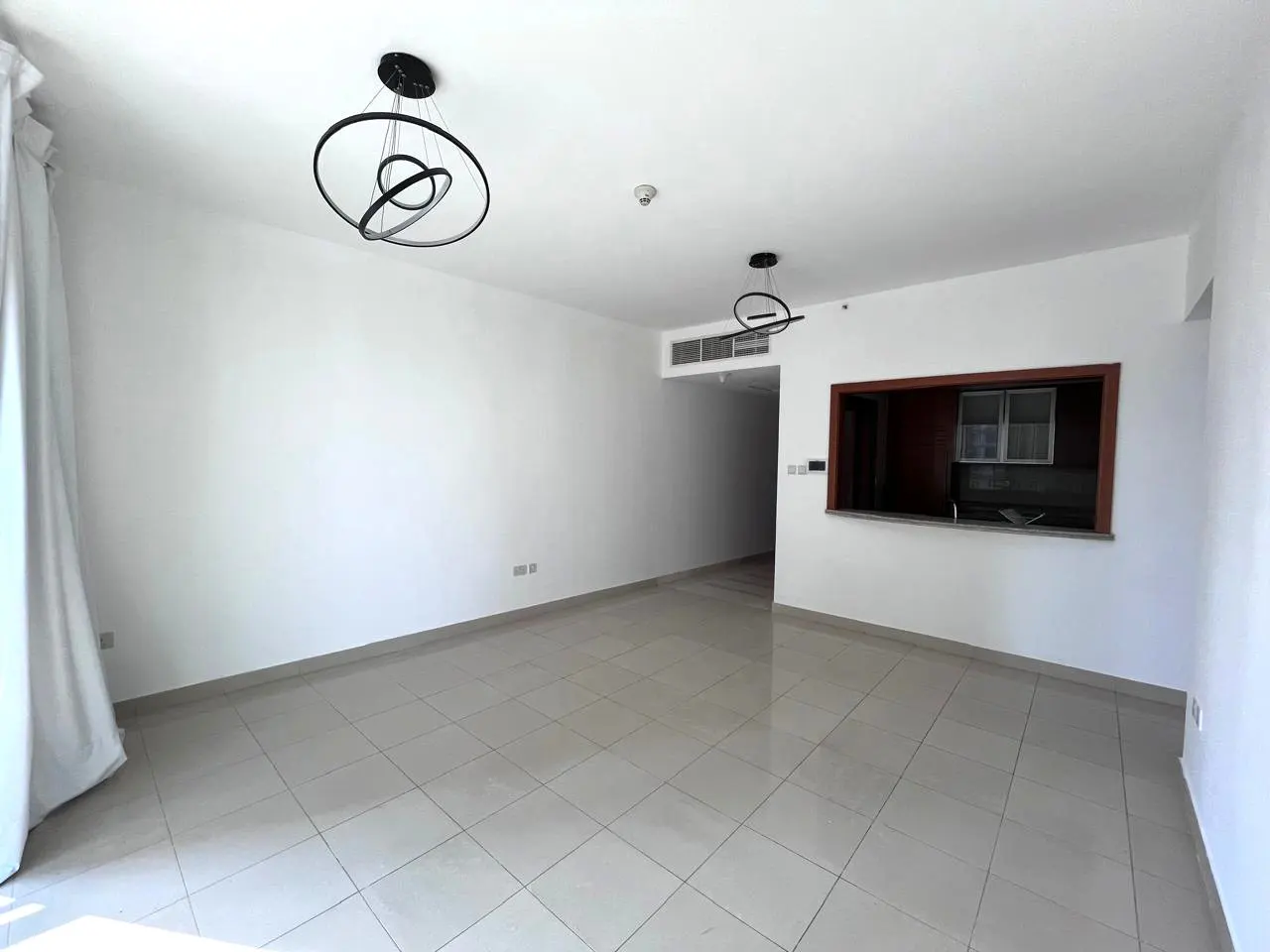 Bright, empty room with white walls, tile floor, and modern black light fixtures. A kitchen pass-through is visible.