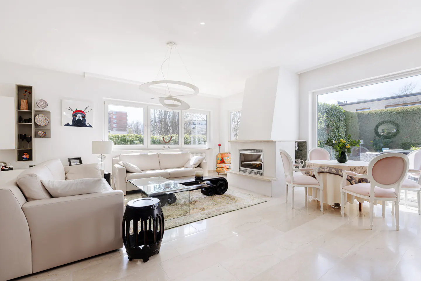 Bright, open living room with white sofas, a glass coffee table, and a fireplace. A dining table sits near a large window overlooking a green garden.