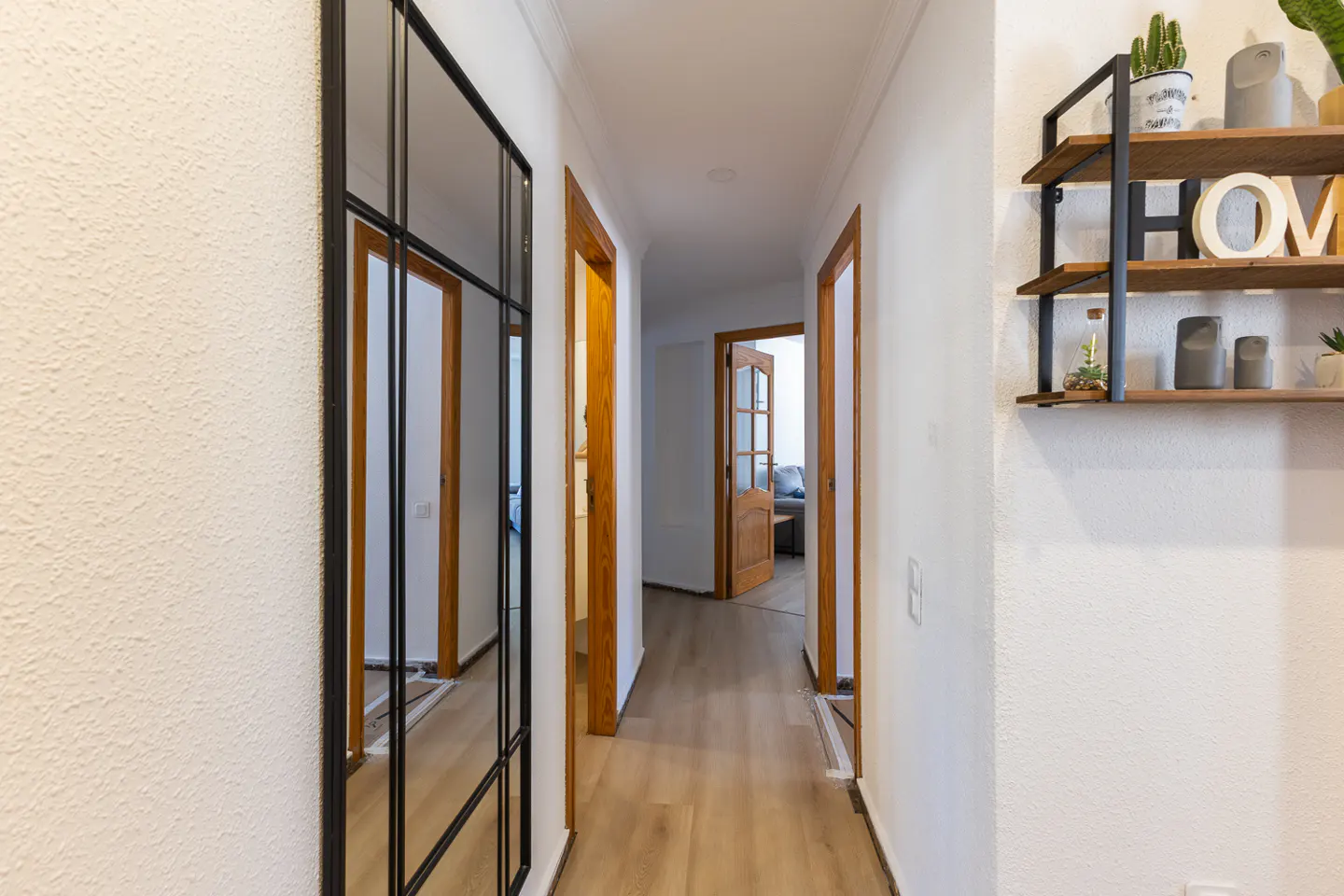 Hallway with light wood floors, white walls, and wood doors. A black framed mirror is on the left wall, and a shelf with decor is on the right.