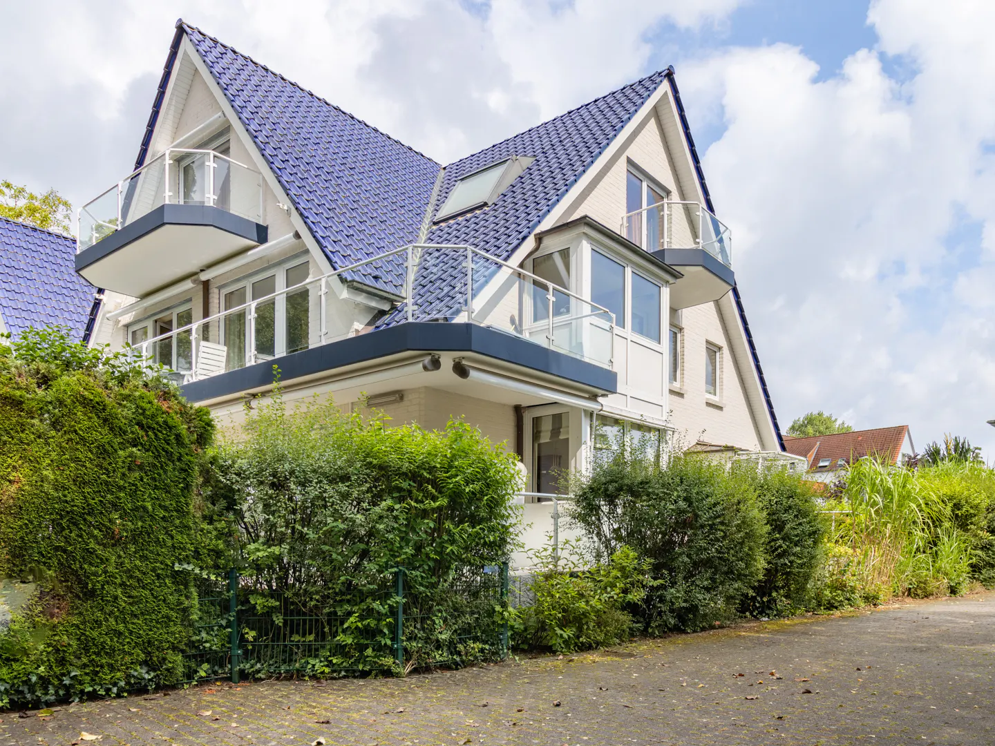 Modern house with a blue tiled roof, multiple balconies, and large windows, surrounded by lush green hedges and a driveway.