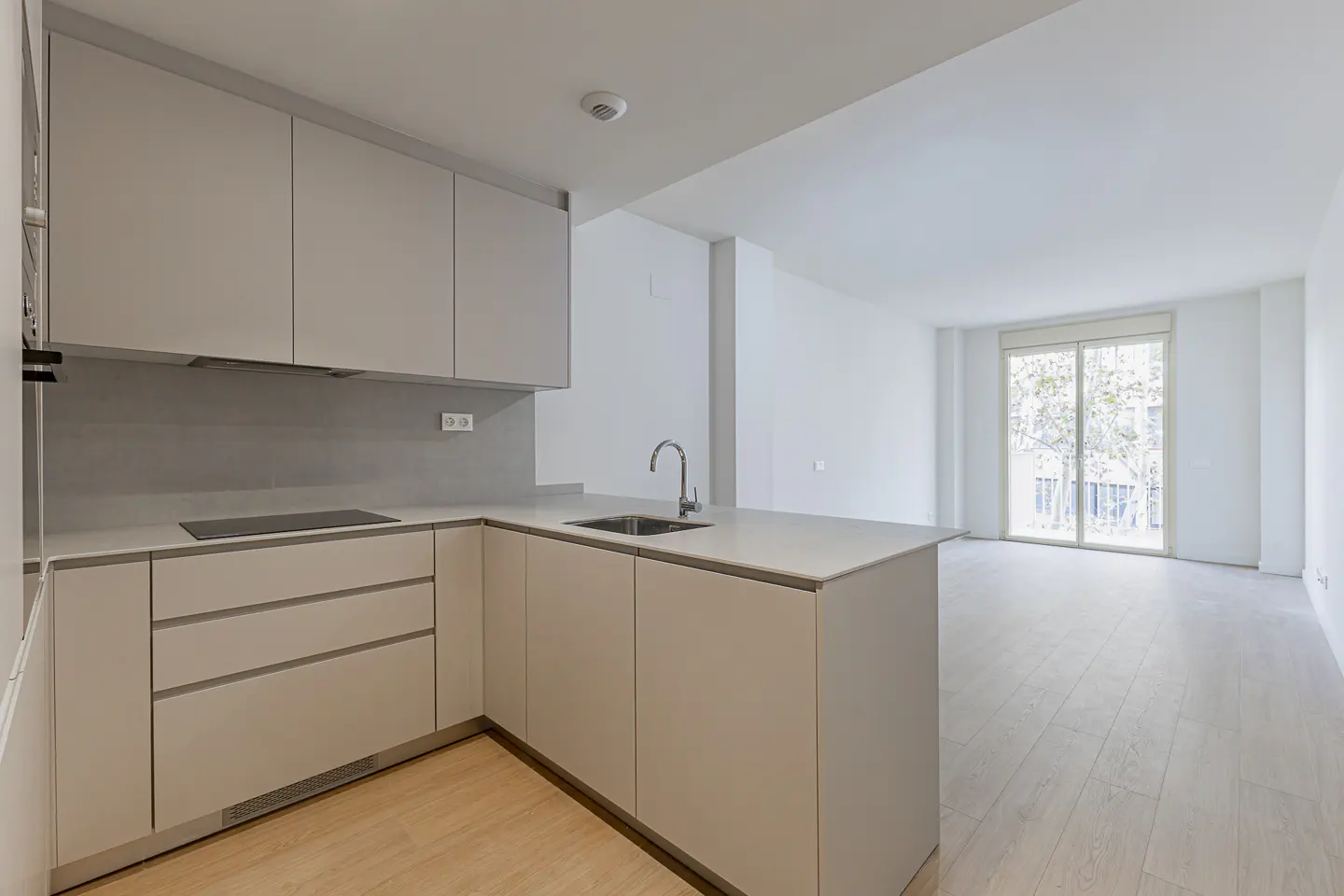 A bright, modern kitchen with light wood floors and white walls. Cabinets are light gray with a stainless steel sink and faucet. A large window is in the background.