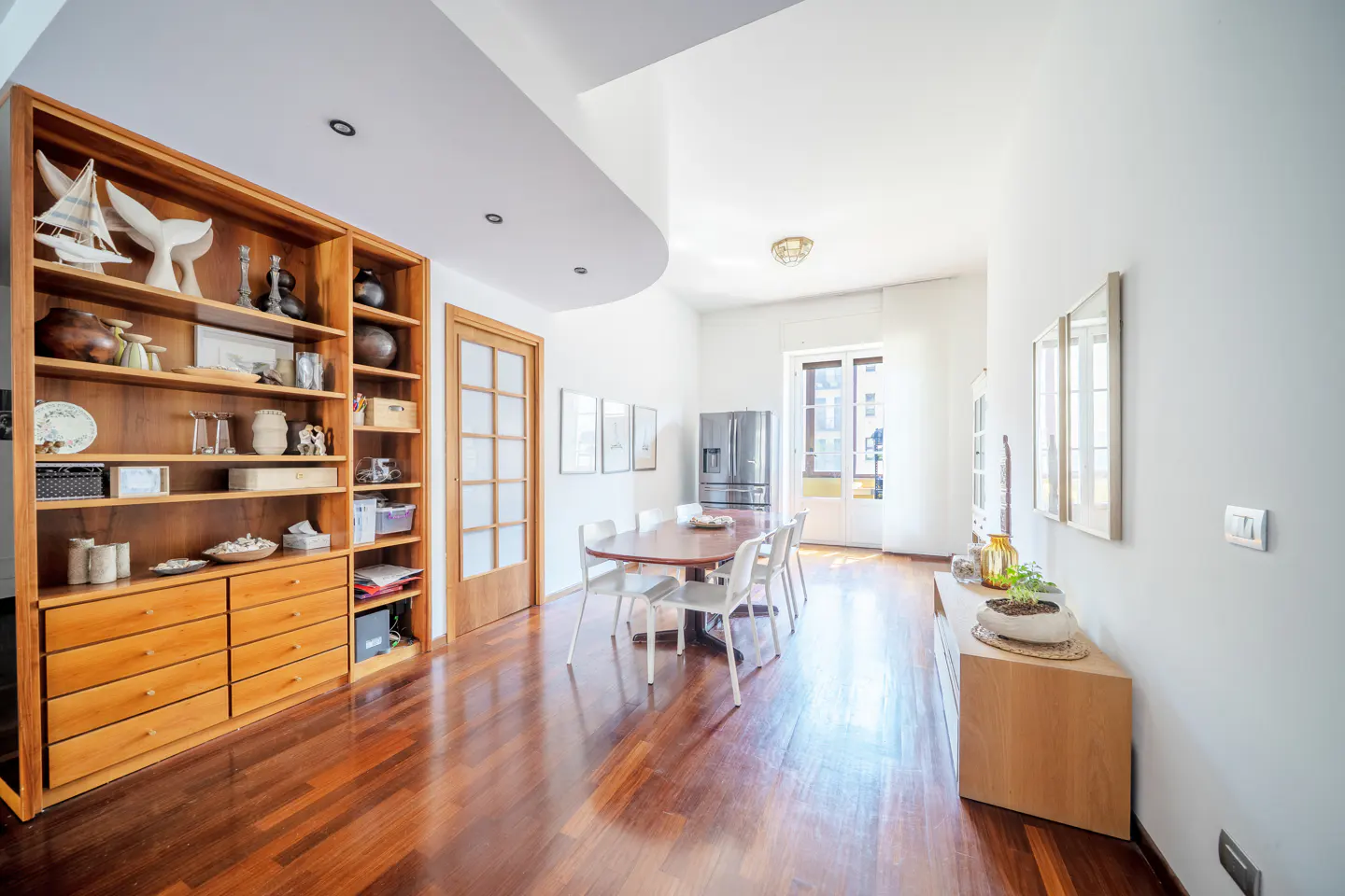 Bright dining room with wood floors, white walls, and a large wooden bookcase filled with decorative items. A dining table with white chairs sits in the center of the room.