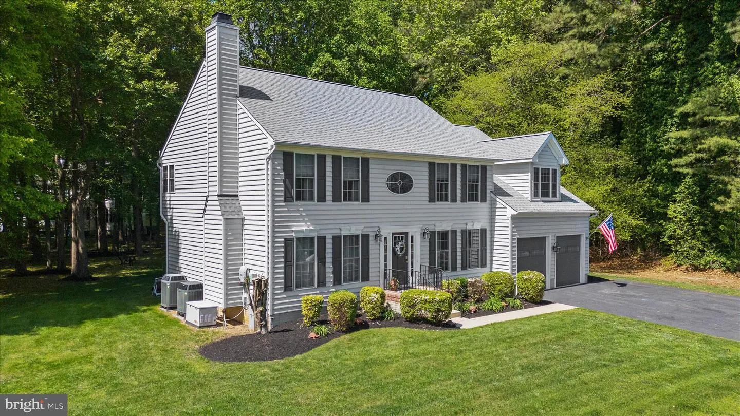 Two-story white house with black shutters, gray roof, and a chimney. Green lawn and trees surround the house. American flag is visible.