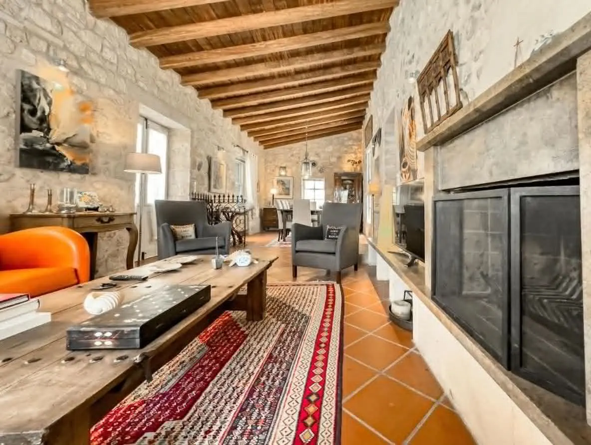 Living room with stone walls, wood beam ceiling, and terracotta tile floor. A large fireplace, gray armchairs, and a long wooden table with a patterned rug are visible.