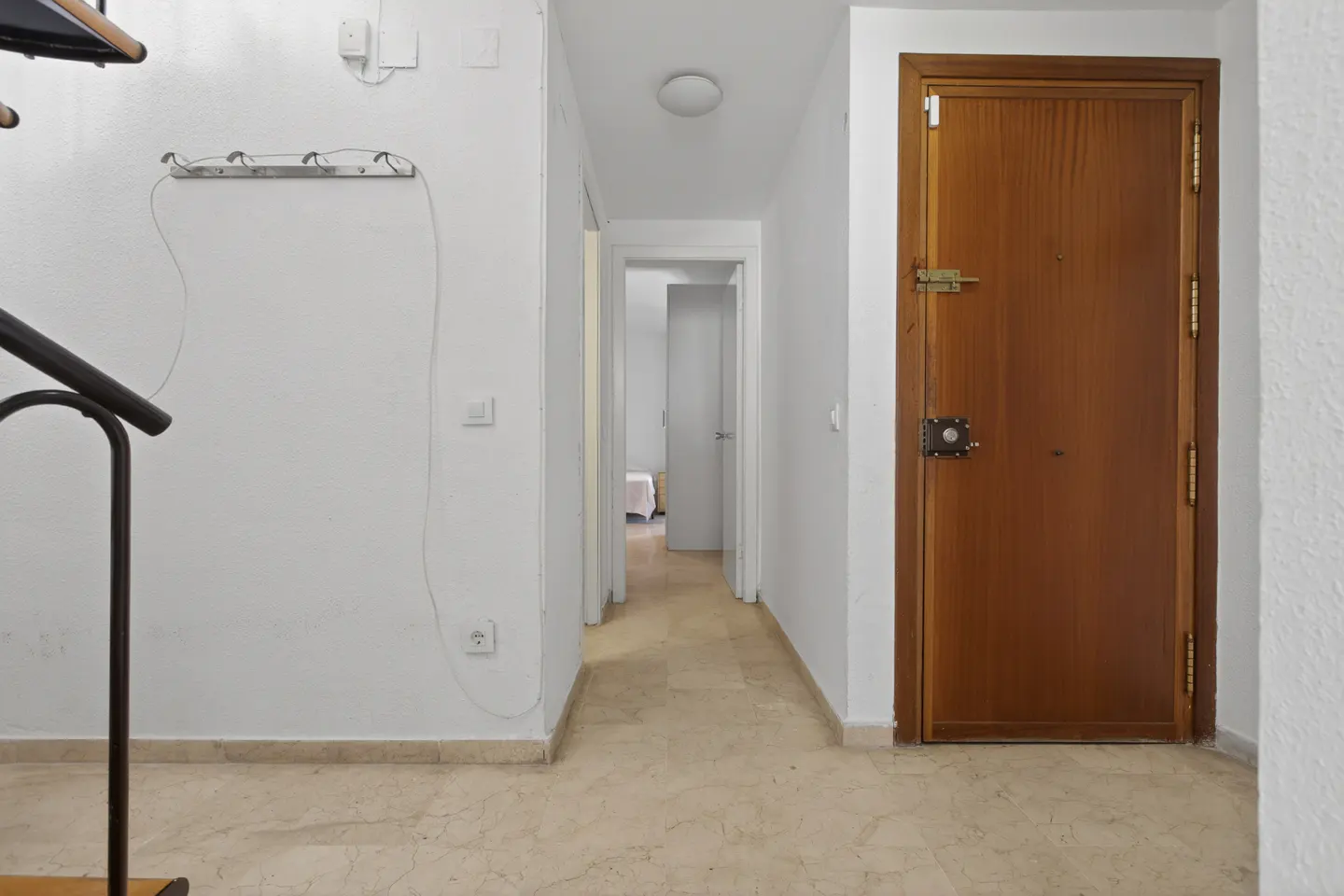 Hallway with a brown door, white walls, and beige marble floor. A coat rack is on the wall, and a glimpse of a bedroom is visible down the hall.