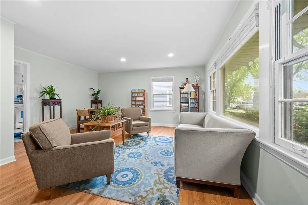 Bright living room with hardwood floors, blue floral rug, two brown armchairs, and a gray sofa near a large window.