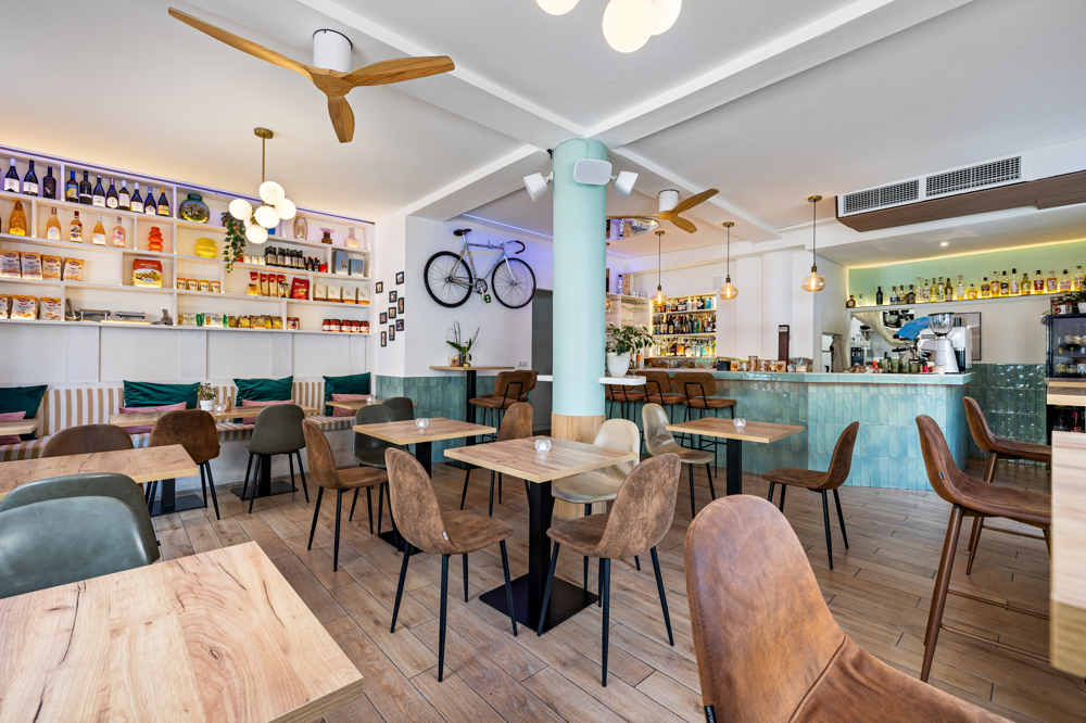 Modern cafe interior with light wood tables, brown chairs, a teal tiled bar, and a bicycle mounted on a white wall.