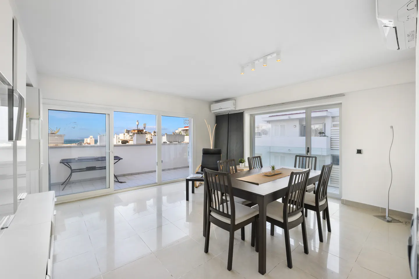 Bright, modern dining room with a dark wood table and chairs. Sliding glass doors lead to a balcony with ocean views. White tile floor.