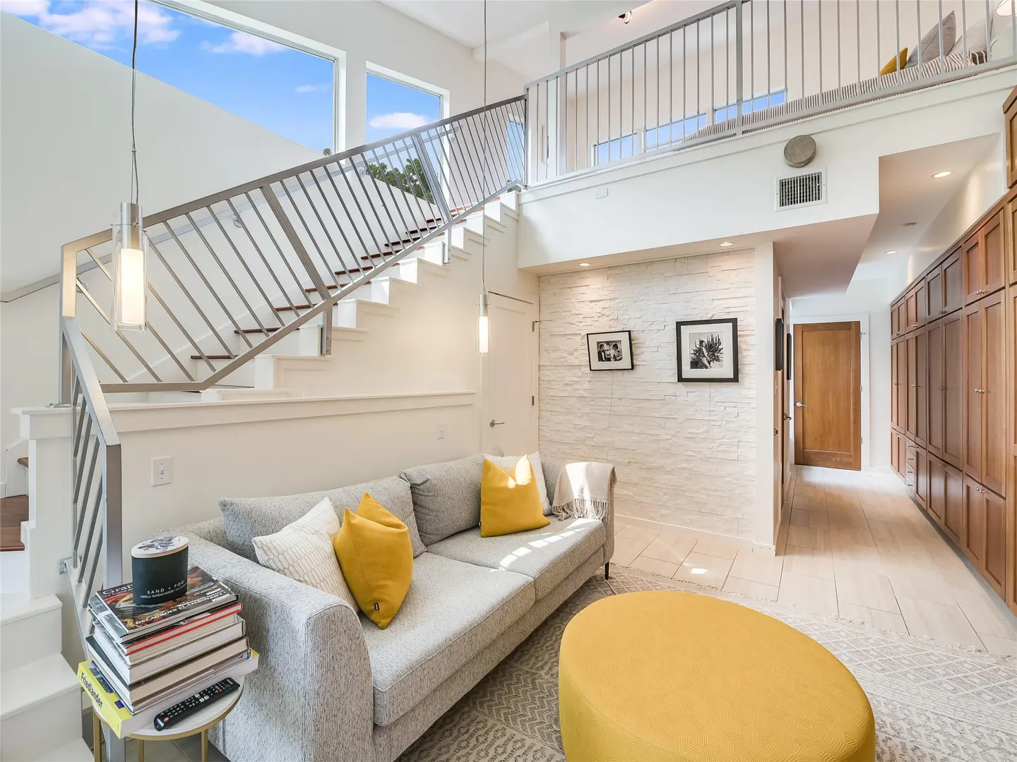 Bright living room with gray sofa, yellow pillows, and ottoman. Stairs lead to a loft area with metal railings. White brick wall with framed art.