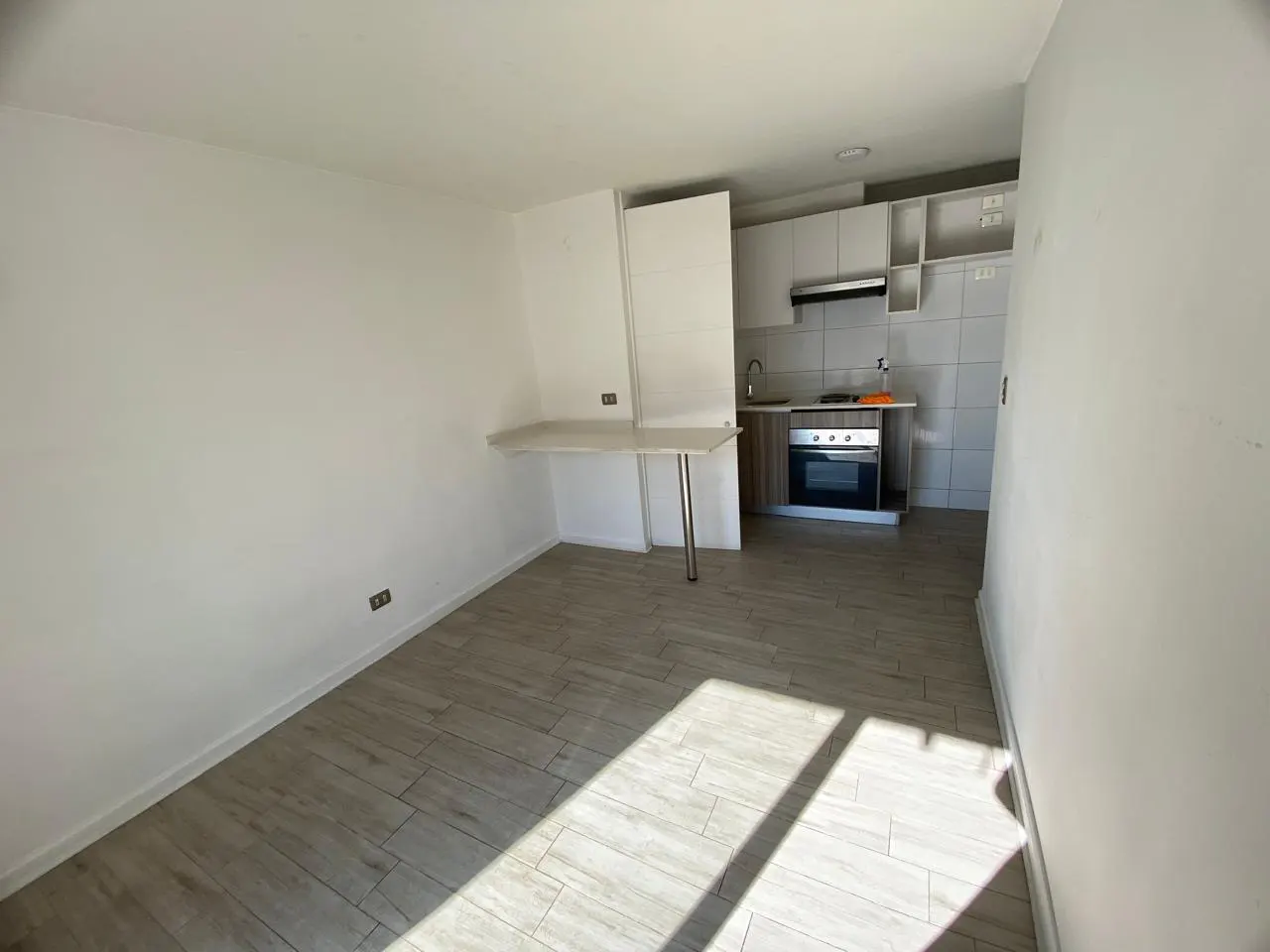 Bright, empty room with wood-look tile floor, white walls, and a small kitchen with white cabinets and stainless steel appliances.
