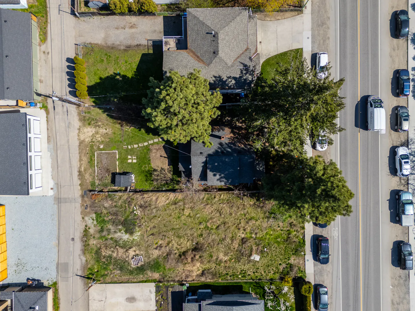 Aerial view of a vacant lot with a small shed, adjacent to a house with a gray roof and a busy street with cars.