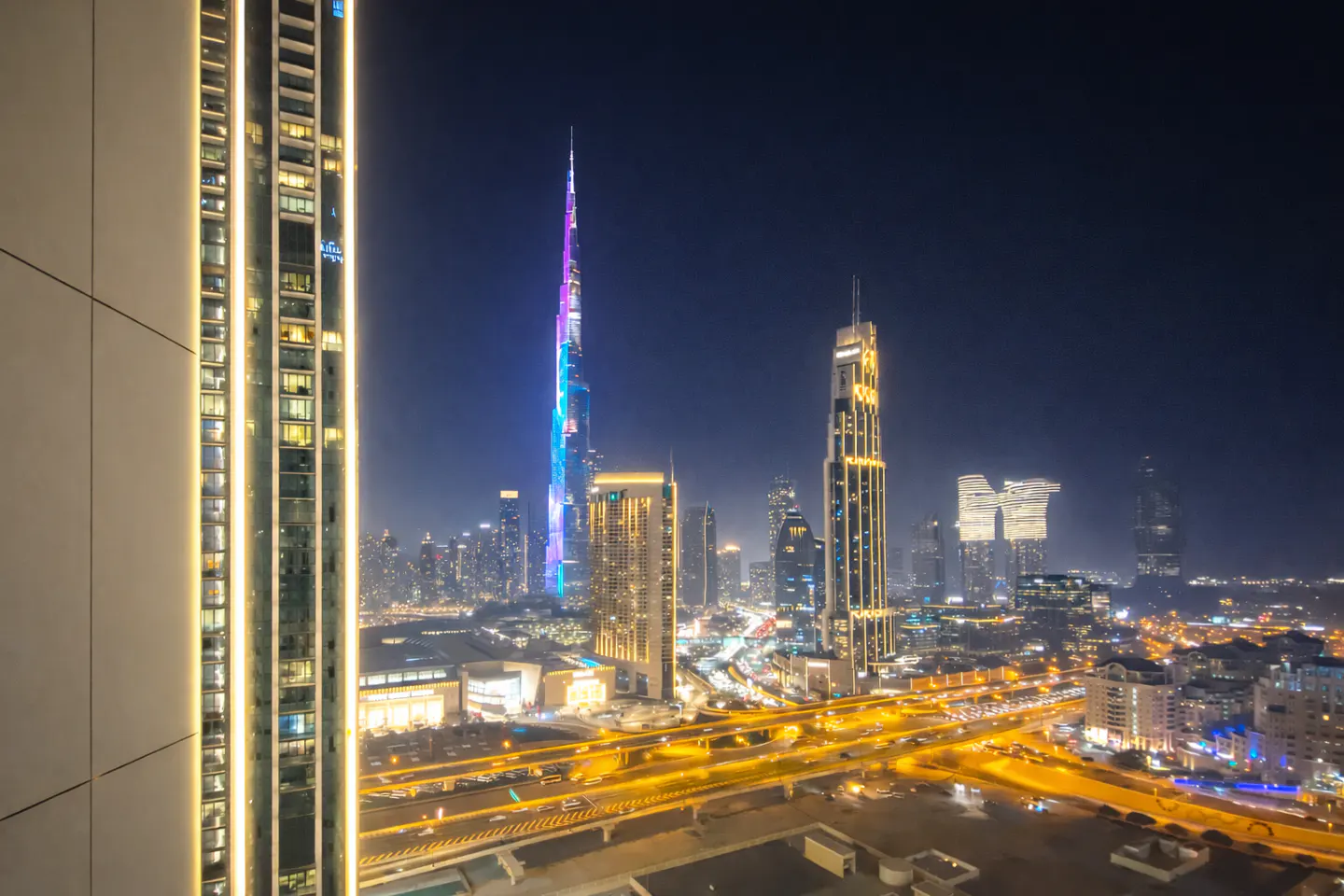 Night view of Dubai skyline featuring Burj Khalifa lit in blue and purple, with bright city lights and highway traffic.