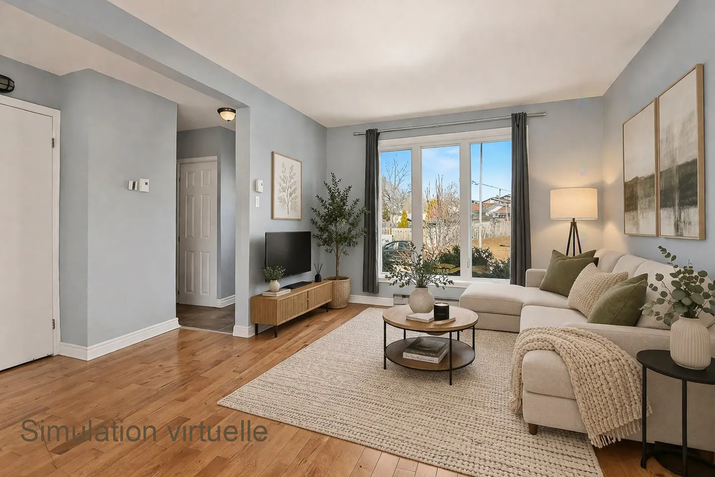 Living room with light blue walls, wood floors, and a large window. A white sectional sofa faces a TV and a round coffee table.