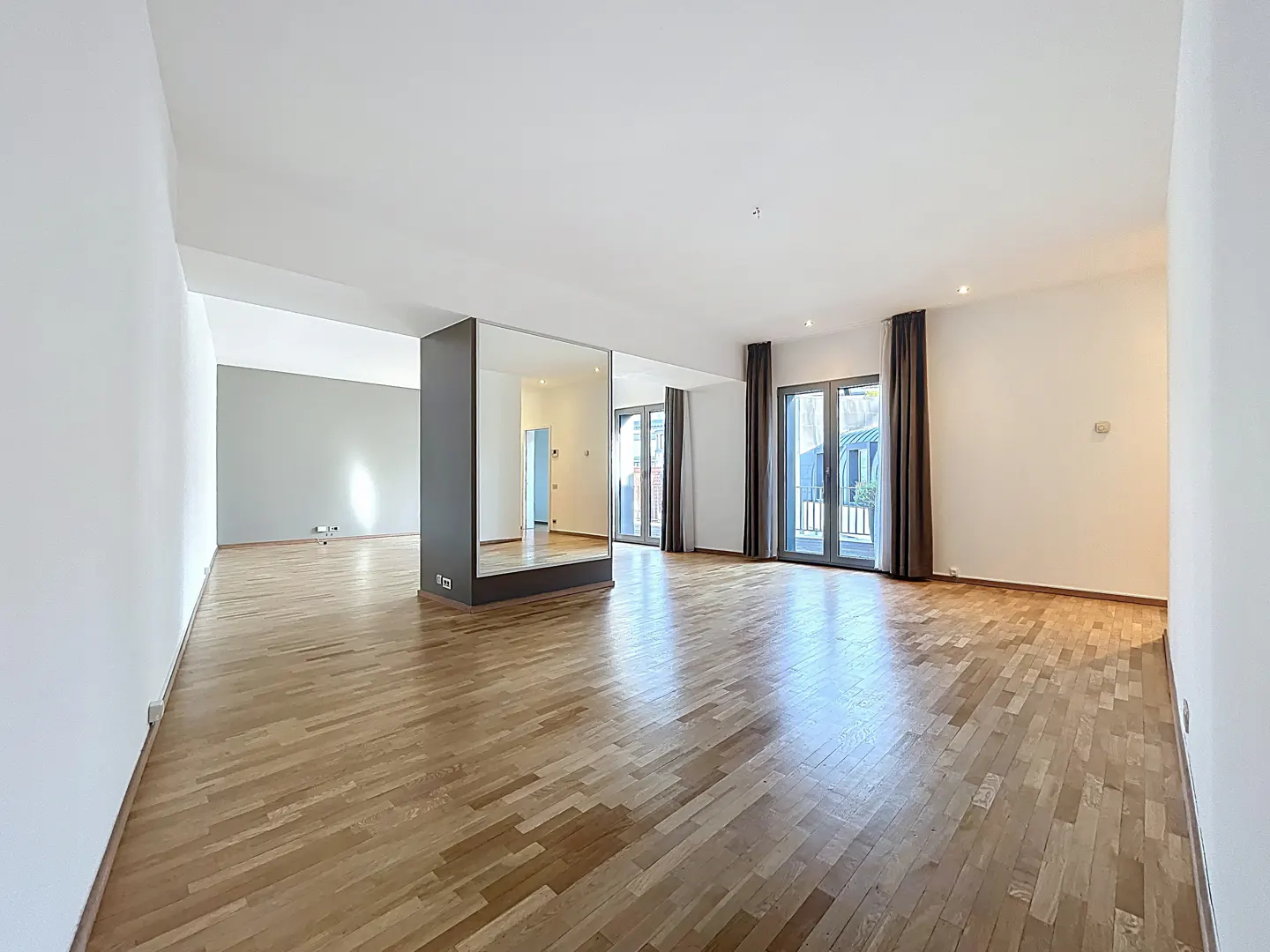 Bright, empty room with hardwood floors, white walls, and a gray accent wall. A large mirror stands near glass doors with brown curtains.