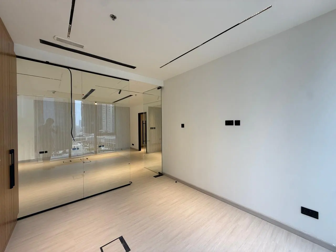 Empty office space with light wood floors, white walls, and glass-walled conference room. Black track lighting on the ceiling.