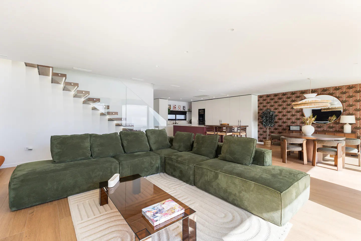 Modern open-plan living room with a large olive green sectional sofa, glass coffee table, and patterned wallpaper.