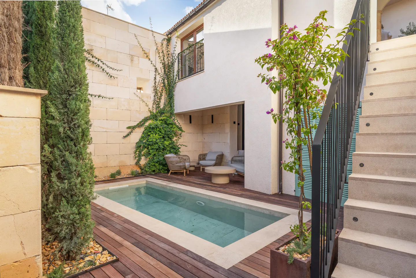 A courtyard with a small pool, wooden deck, and stone walls. Seating area with chairs and a table. Stairs with black railing on the right.