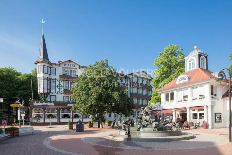 Exterior view of a European town square with a fountain, hotel, and shops on a sunny day.