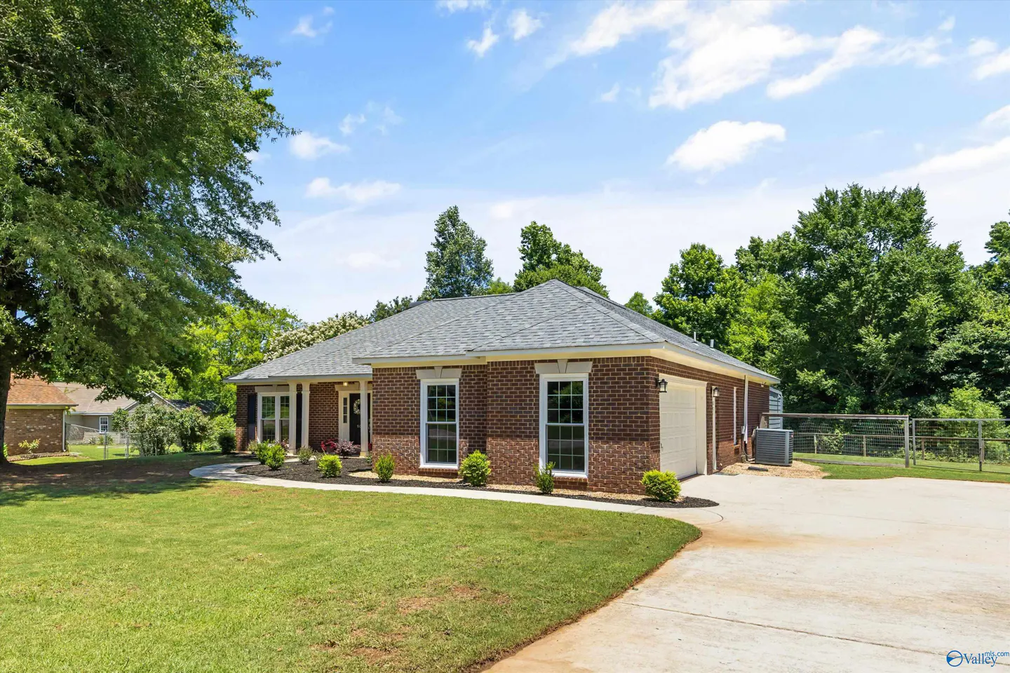 Single-story red brick house with a gray shingled roof, concrete driveway, and a large green lawn under a blue sky.