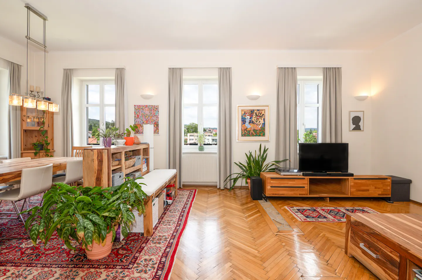 Bright living room with herringbone wood floors, a dining area, a TV console, and large windows with grey curtains.