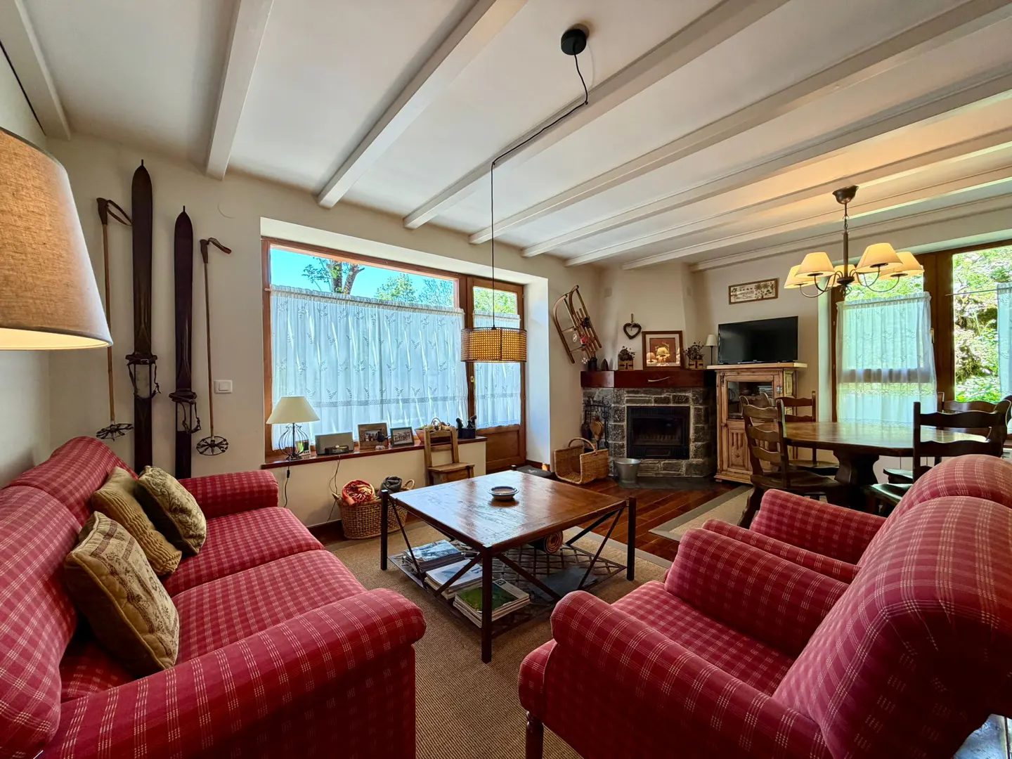 Living room with red plaid sofas, wooden table, fireplace, and skis on the wall. Natural light through the window.