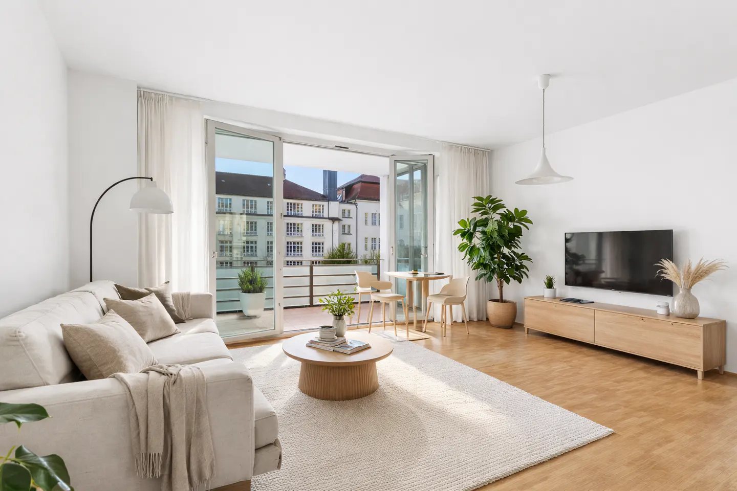 Bright living room with white sofa, round wood table, and open balcony doors. A TV and potted plant sit against the white wall.