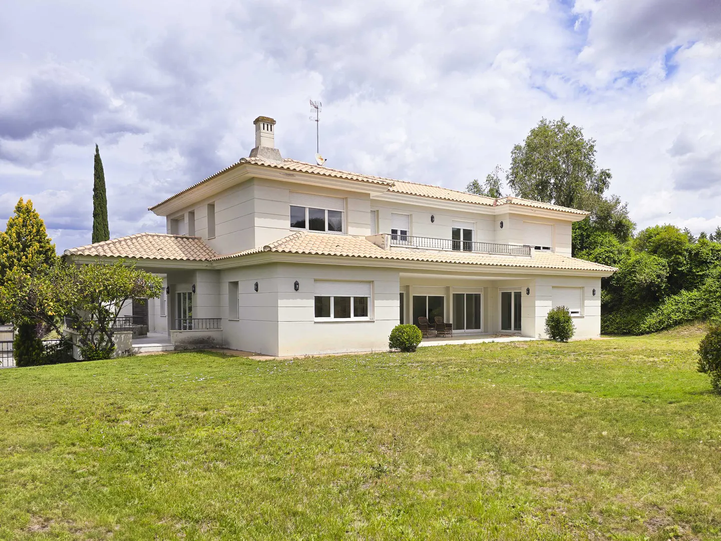 Two-story beige house with a tan tile roof, chimney, and balcony, surrounded by a green lawn and trees under a cloudy sky.