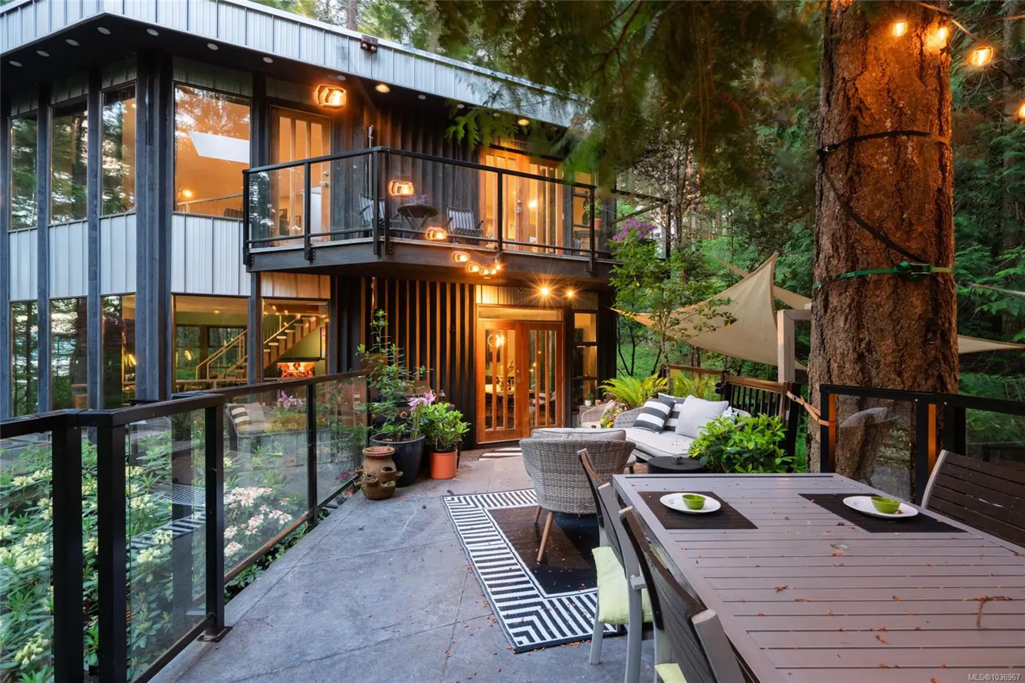 A modern two-story house with a deck featuring a dining table, chairs, and outdoor seating area. String lights illuminate the scene.