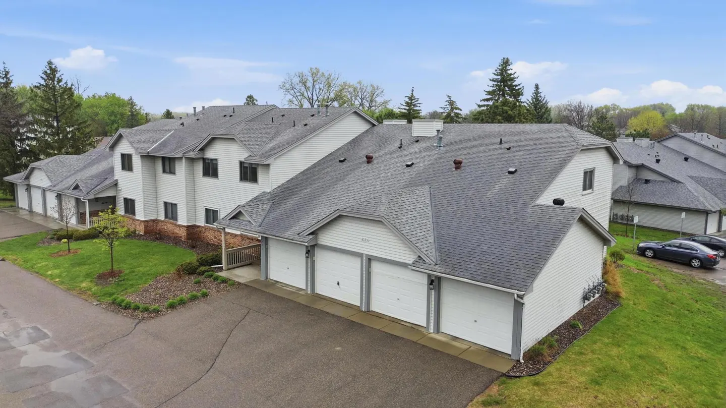 Aerial view of light gray townhouses with gray roofs and white garage doors, surrounded by green grass and trees.