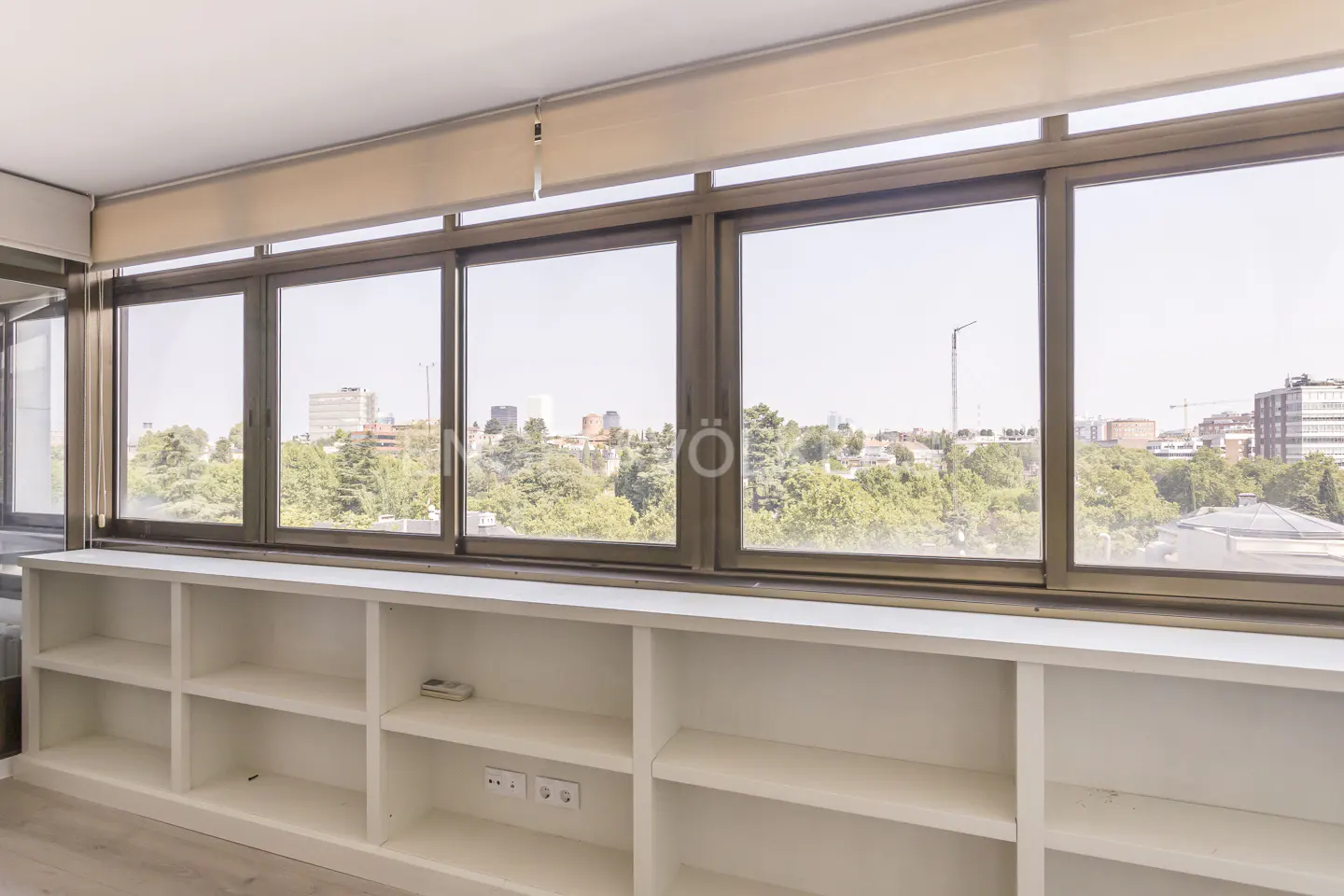 Bright room with large windows overlooking a cityscape. White built-in shelves line the wall below the windows.