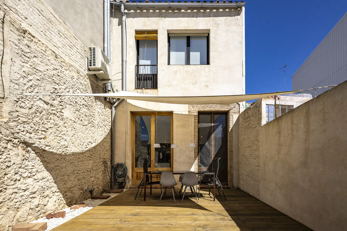 Outdoor patio with wooden deck, table, and chairs, surrounded by textured walls and a sunshade.